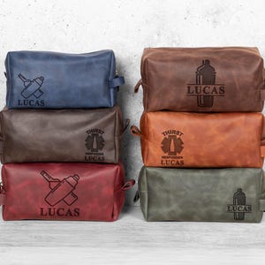 May include: A stack of six leather toiletry bags in various colors: blue, brown, red, tan, and green. Each bag features a unique design, including a cocktail shaker, a medical symbol, and the name "LUCAS".