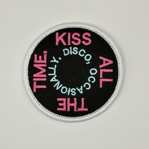 May include: A circular embroidered patch with a black background and a white stitched border. The text reads KISS ALL THE TIME in pink and OCCASIONALLY DISCO in light blue, arranged in a circular pattern around the center of the patch.