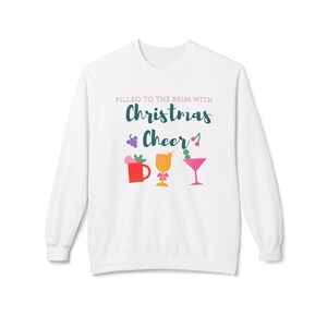 May include: White sweatshirt with the text "FILLED TO THE BRIM WITH Christmas Cheer" in green and pink. The design includes colorful illustrations of a mug, a cocktail glass, and a martini glass, perfect for the holiday season.