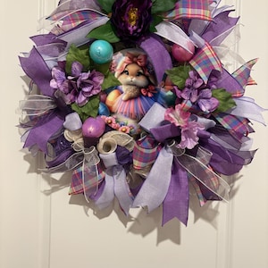 May include: Purple and pink Easter wreath with a bunny holding Easter eggs. The wreath is decorated with plaid ribbon, mesh, and flowers.