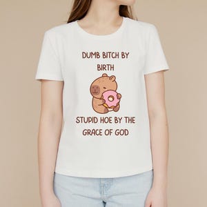 May include: White t-shirt with a cartoon capybara holding a pink donut. The text on the shirt reads "DUMB BITCH BY BIRTH STUPID HOE BY THE GRACE OF GOD" in brown. The t-shirt has a crew neck and short sleeves.