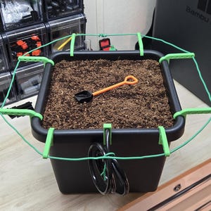 Könnte beinhalten: Ein schwarzer quadratischer Pflanzkübel mit Erde, mit einer kleinen orangefarbenen Schaufel. Eine grüne Stützstruktur umgibt den Pflanzkübel. Ein kleiner grüner Behälter mit Erde befindet sich links. Die Szene spielt auf einer Holzoberfläche.