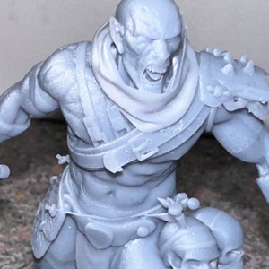 Puede incluir: Una miniatura gris impresa en 3D de una figura humanoide con una expresión feroz. La figura lleva una capa con capucha y un cinturón con una hebilla. La figura sostiene una calavera en su mano derecha y tiene una calavera atada a su cinturón.