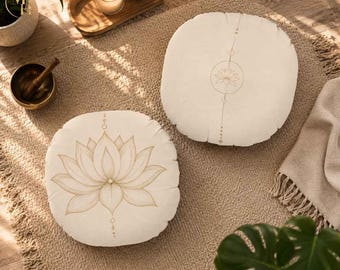 May include: Two round, off-white floor cushions featuring a delicate, light brown line art illustration of a lotus flower. The design includes vertical geometric accents with small circles and dots centered above and below the lotus motif.