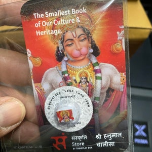 Puede incluir: Un pequeño libro rojo, blanco y dorado con una imagen de una deidad hindú en la portada. El libro mide menos de 2,5 cm y está hecho a mano. El texto de la portada dice "The Smallest Book of Our Culture & Heritage."  "Sanskriti Store" y "Shri Hanuman Chalisa" también están impresos en la portada.