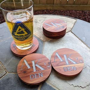 Puede incluir: Cuatro posavasos redondos de madera con el texto "AK's Den" grabado en letras plateadas. Un vaso de cerveza se apoya en uno de los posavasos.