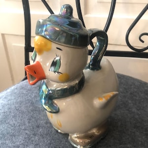 Può includere: Teiera in ceramica a forma di anatra con finitura perlata. La teiera presenta un cappello e una sciarpa blu, un becco arancione e dettagli gialli. Anche il manico e il beccuccio sono blu.