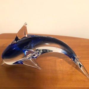 Puede incluir: Una escultura de delfín de cristal con un diseño elegante y curvado. El delfín presenta un degradado de vidrio azul y transparente, con una aleta dorsal prominente. La escultura es un artículo decorativo.