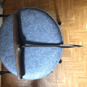 May include: A round, gray upholstered stool with a dark wooden tool resting on top. The tool has a long, slender metal shaft and a wooden handle. The stool is on a wooden floor.