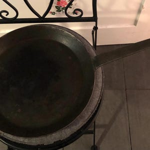 Puede incluir: Una sartén grande de metal oscuro con un mango largo, probablemente para cocinar. La sartén redonda tiene una superficie de cocción amplia y plana. Descansa sobre un soporte de metal decorativo, lo que sugiere un estilo vintage o rústico. Diámetro aproximado de 40 cm.