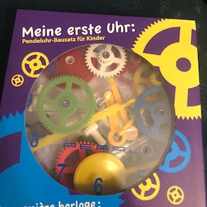 May include: A boxed clock-making kit for children. The kit includes colorful plastic gears, clock hands, and a pendulum. The box has the text "Meine erste Uhr: Pendeluhu-Bausatz für Kinder" in white letters on a purple background.