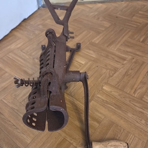 Könnte beinhalten: Ein antiker, rostiger Metall-Fleischwolf mit Holzgriff. Der Wolf hat eine Kurbel, einen Trichter und eine Klemme zur Befestigung. Der Griff ist aus hellfarbenem Holz. Der Wolf wird wahrscheinlich zum Zerkleinern von Fleisch oder Gemüse verwendet.