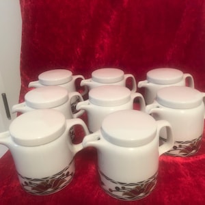 May include: Set of eight white ceramic creamer pitchers with brown and gold floral designs. The pitchers have lids and are arranged in a semi-circle on a red velvet surface.