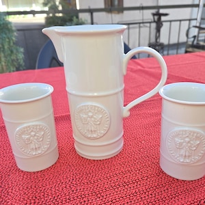 Puede incluir: Una jarra de cerámica blanca y dos vasos a juego, cada uno con un sello circular en relieve que representa una figura sosteniendo un bastón. El conjunto tiene un acabado liso y brillante, forma cilíndrica y un asa curva en la jarra.