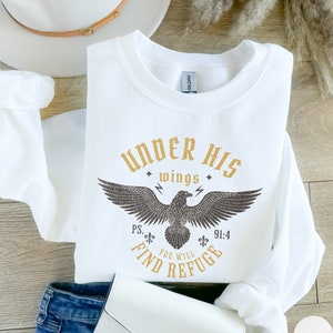 May include: White sweatshirt with a gold graphic of an eagle with the text "Under His Wings You Will Find Refuge" and the scripture reference "Ps. 91:4".