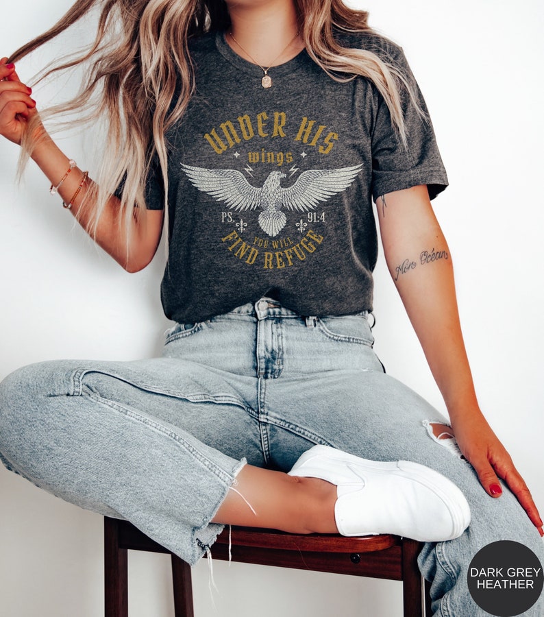 May include: A dark grey heather t-shirt with a white eagle graphic and the text "Under His Wings You Will Find Refuge" in a vintage style font. The text "PS 91:4" is also included.