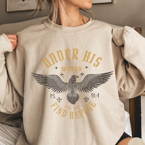 May include: A beige sweatshirt with a graphic print of an eagle with the text "Under His Wings" and "Ps. 91:4 You Will Find Refuge".