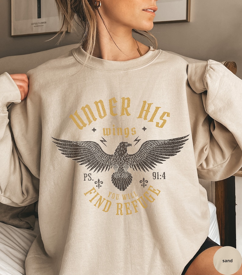 May include: A beige sweatshirt with a graphic print of an eagle with the text "Under His Wings" and "Ps. 91:4 You Will Find Refuge".