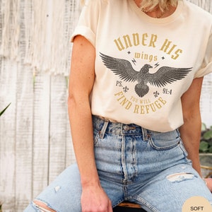 May include: A cream colored t-shirt with a graphic of an eagle with the text "Under His Wings You Will Find Refuge" and the scripture reference "Ps. 91:4".