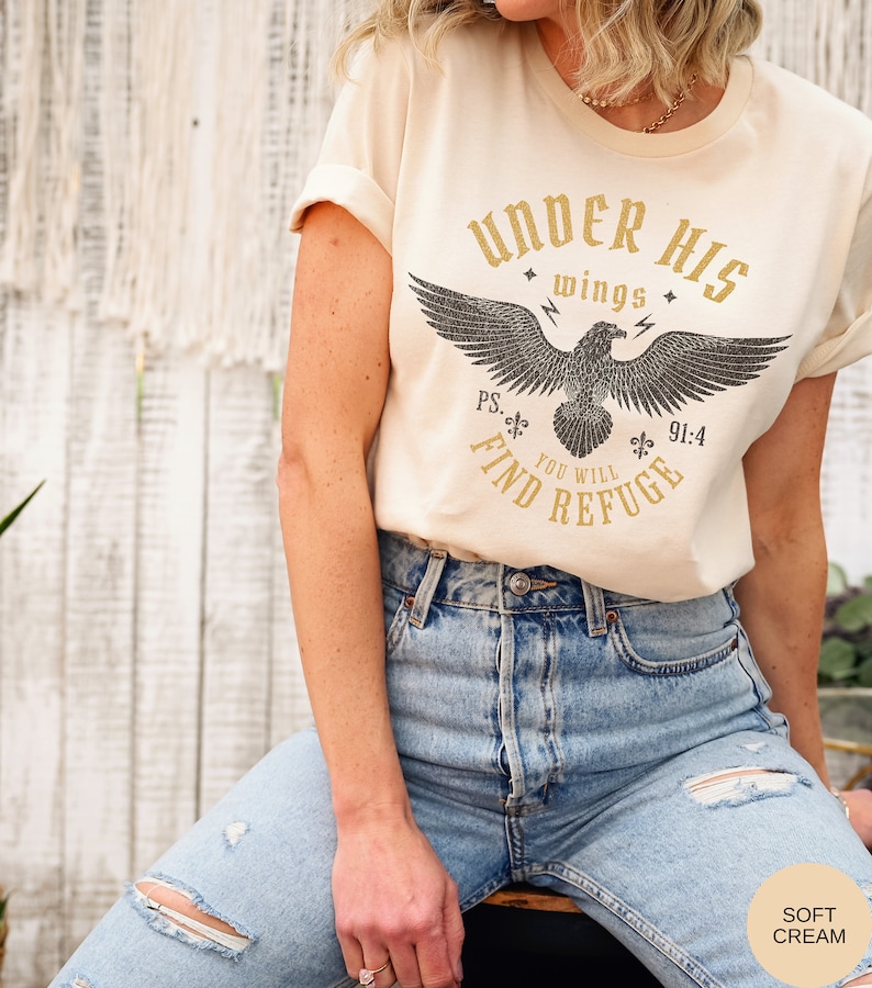 May include: A cream colored t-shirt with a graphic of an eagle with the text "Under His Wings You Will Find Refuge" and the scripture reference "Ps. 91:4".