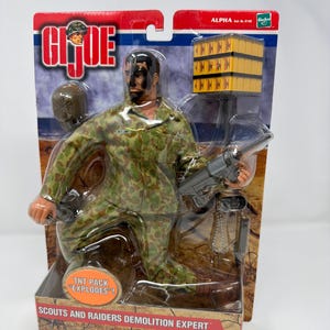 Może przedstawiać: Figurka G.I. Joe w kamuflażu, trzymająca karabin maszynowy. Figurka jest zapakowana z pakietem TNT i oznaczona jako "Scouts and Raiders Demolition Expert". Opakowanie zawiera logo G.I. Joe i ostrzeżenie o małych częściach.