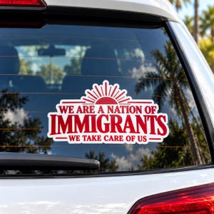 May include: A white vinyl decal applied to a car window featuring red text that reads WE ARE A NATION OF IMMIGRANTS WE TAKE CARE OF US with a red sunburst graphic above the text. The design is centered on the rear glass of the vehicle.