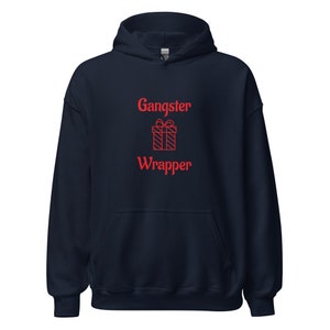 Puede incluir: Sudadera con capucha azul marino con las palabras "Gangster Wrapper" en rojo y un gráfico de caja de regalo roja. La sudadera tiene un bolsillo delantero y una capucha con cordón.