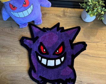 Puede incluir: Alfombra morada y negra con una imagen de dibujos animados de un fantasma con ojos rojos y una amplia sonrisa. La alfombra está sobre un suelo de madera. El texto "TOUGH MUDDER 10K" está en una diadema amarilla que lleva un fantasma de peluche morado.