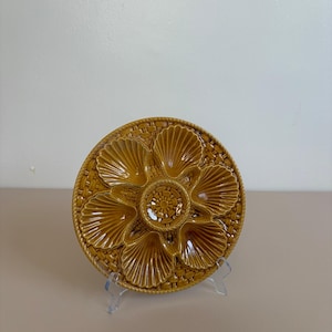 May include: A round, mustard-yellow ceramic oyster plate featuring six shell-shaped indentations arranged in a circular pattern around a central textured medallion. The background of the plate has a woven basket-weave pattern with a rope-style border.