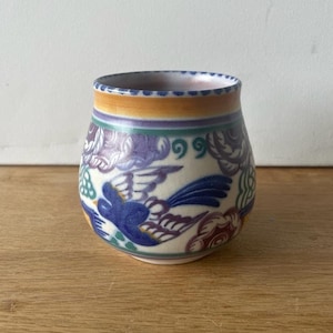 May include: A small, white ceramic vase with a rounded body and a wide mouth. The vase is decorated with a colorful floral pattern and a blue bird design.