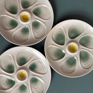 May include: Three round ceramic oyster plates with a light gray finish. Each plate features six shell-shaped indentations with green gradient centers and a central circular yellow well for condiments, arranged in a radial pattern on the surface.
