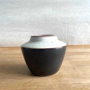 May include: A small, two-toned ceramic vase. The bottom half is a dark, matte black, while the top half is a textured, off-white. A thin, brown band accents the rim. The vase sits on a light-colored wooden surface.