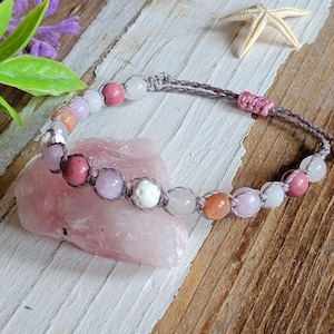 May include: A gray macrame bracelet with pink, white, and orange beads. The bracelet is on a pink and white surface.