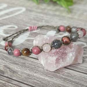 May include: A beaded bracelet featuring a variety of colorful gemstones, including pink, brown, and gray beads. The bracelet is strung on a gray cord and rests on a pink crystal. The bracelet is a piece of jewelry.