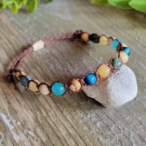 May include: A brown cord bracelet with a variety of gemstone beads, including blue, green, yellow, and white. The bracelet is tied with a knot and is resting on a white rock.