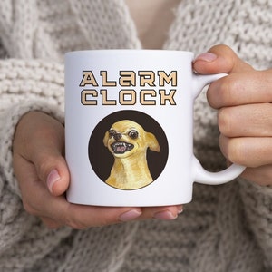 May include: White ceramic mug with a black circle containing a cartoon chihuahua with a grumpy expression. The text "ALARM CLOCK" is printed in black with a gold outline.