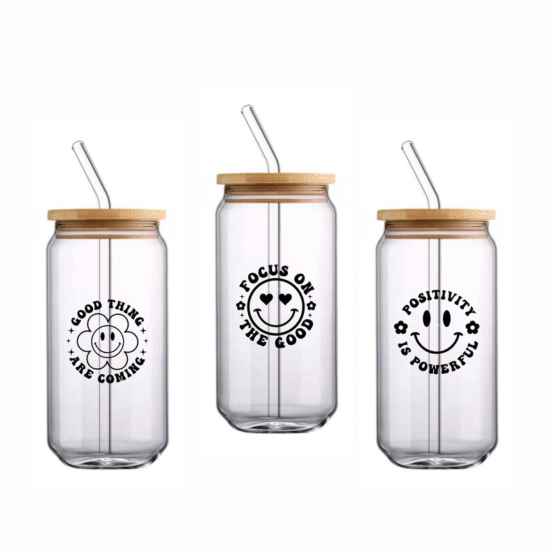 May include: Three clear glass tumblers with bamboo lids and glass straws. Each tumbler has a different black graphic: a smiling flower, a smiley face with hearts, and a smiley face with flowers. The text on the tumblers reads "Good things are coming", "Focus on the good", and "Positivity is powerful".