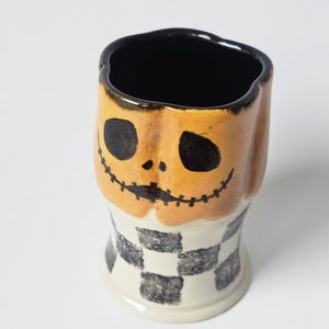 May include: A ceramic mug featuring a sculpted pumpkin head with a hand-painted black face, including hollow eyes and a stitched mouth. The base of the mug is cylindrical and decorated with a black and white checkered pattern, with a black interior.