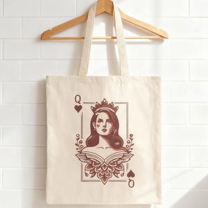 May include: Cream-colored tote bag with a playing card design. The design features a woman wearing a crown, framed within a playing card border, with the letter "Q" and a heart symbol at the corners. The bag hangs from a wooden hanger.