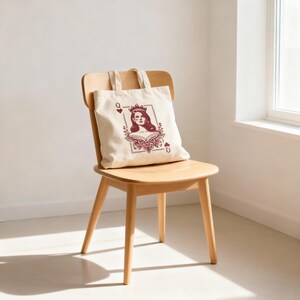 May include: A cream-colored tote bag featuring a maroon illustration of a queen card. The design includes a woman's portrait, a crown, and floral elements. The bag is sitting on a wooden chair.