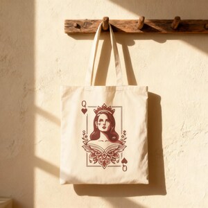 May include: Cream-colored tote bag featuring a Queen of Hearts design. The illustration depicts a woman with a crown, framed by floral elements and the suit symbols. The bag hangs from a wooden rack against a light-colored wall.