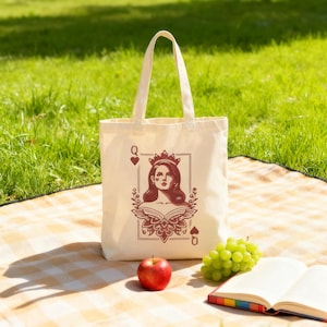May include: A cream-colored tote bag with a graphic of a woman wearing a crown, framed by playing card symbols. The bag is on a picnic blanket with an apple, grapes, and an open book.