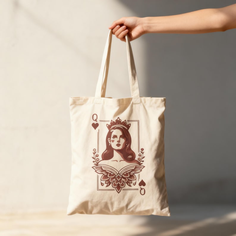 May include: Cream-colored tote bag featuring a Queen of Hearts playing card design. The illustration is in shades of brown and depicts a woman with a crown and floral accents. The bag has long handles for carrying.