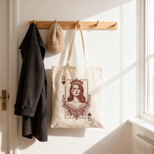 May include: Cream-colored tote bag featuring a Queen of Hearts design in burgundy. The illustration includes a woman wearing a crown, framed by floral elements and card suit symbols. A dark gray coat and tan beanie hang on a wooden rack.