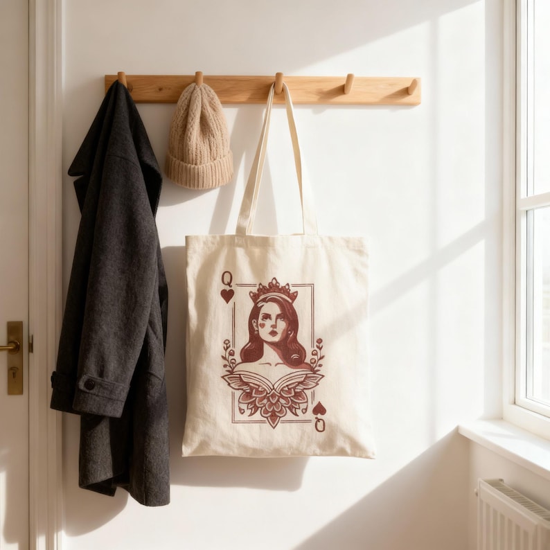 May include: Cream-colored tote bag featuring a Queen of Hearts design in burgundy. The illustration includes a woman wearing a crown, framed by floral elements and card suit symbols. A dark gray coat and tan beanie hang on a wooden rack.