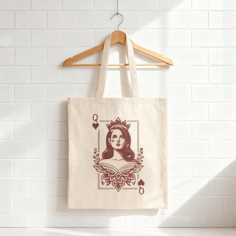 May include: Cream-colored tote bag with a playing card design. The design features a woman wearing a crown, framed within a playing card border, with the letter "Q" and a heart symbol at the corners. The bag hangs from a wooden hanger.