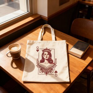 May include: Cream-colored tote bag featuring a Queen of Hearts playing card design. The illustration includes a woman with a crown, surrounded by floral elements, and the heart suit symbols. The bag has a simple, rectangular shape with two handles.
