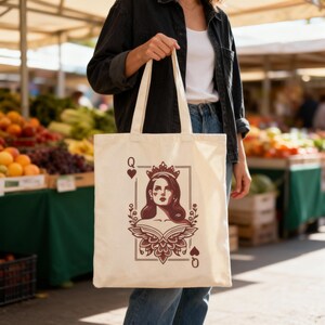 May include: Cream-colored tote bag featuring a Queen of Hearts design. The illustration depicts a woman wearing a crown, framed by a rectangular border and floral accents. The bag has long handles and is held in front of a market background.