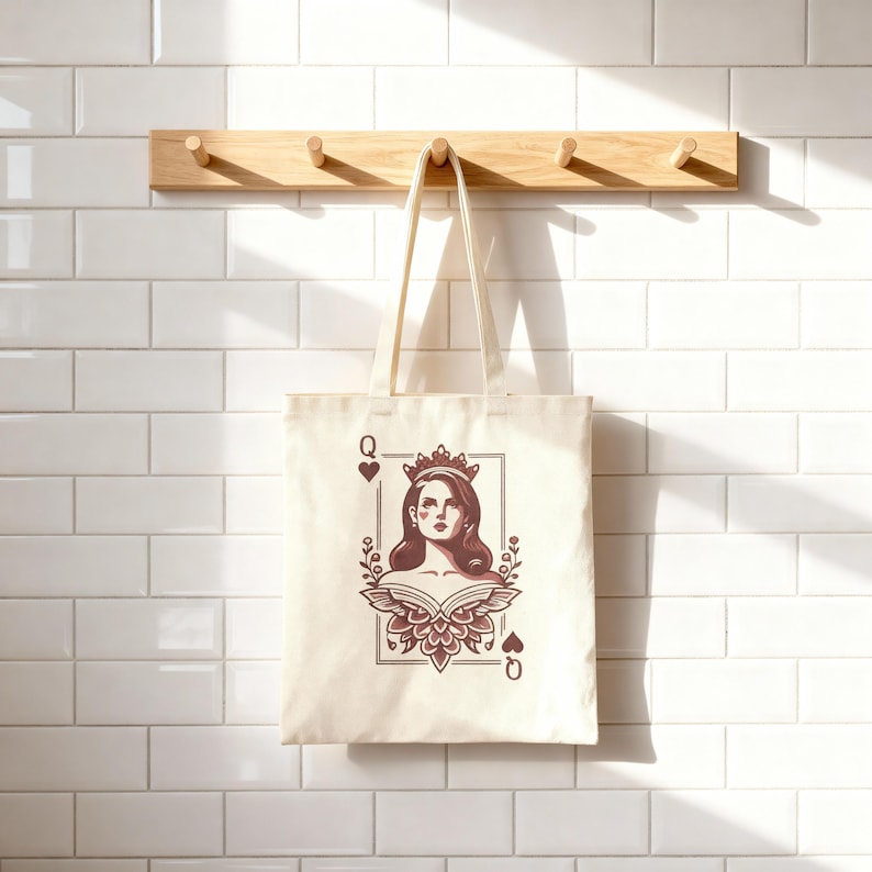 May include: Cream-colored tote bag featuring a Queen of Hearts design. The illustration shows a woman with a crown, framed by floral elements and playing card symbols. The bag hangs from a wooden rack against a white tiled wall.