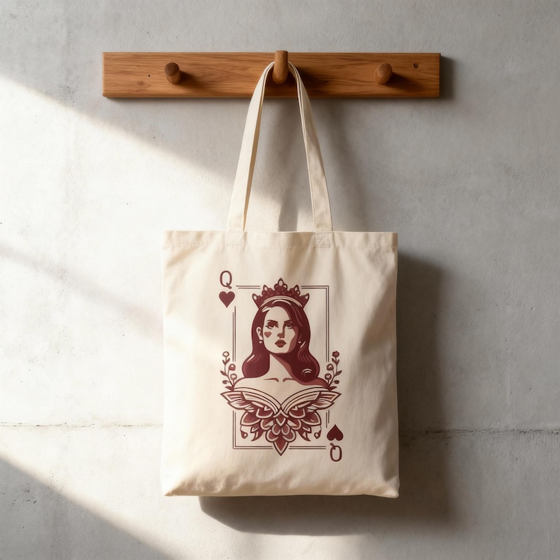 May include: Cream-colored tote bag with a maroon illustration of a queen from a deck of cards. The queen is depicted with a crown and floral accents. The bag hangs from a wooden rack on a gray wall.